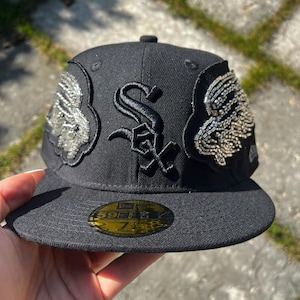 Puede incluir: Una gorra de béisbol negra con un logotipo de los Chicago White Sox bordado en relieve en el centro. La gorra está adornada con dos parches simétricos en los laterales, decorados con cuentas metálicas plateadas. Una pegatina dorada y negra está en la visera.