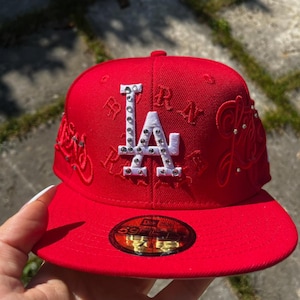 Könnte beinhalten: Eine leuchtend rote Baseballkappe mit einem weiß gestickten LA-Logo, das mit klaren Strasssteinen verziert ist. Die Kappe zeigt zusätzliche rote Ton-in-Ton-Stickereien und verstreute Strasssteine an den Seiten sowie einen 59FIFTY-Aufkleber auf dem flachen Schirm.