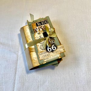 Può includere: Un piccolo libro fatto a mano con un'estetica vintage e usurata. È rilegato con carta marrone e decorato con vari elementi effimeri, tra cui un nastro dorato, un'etichetta metallica con il numero "66" e un quadrante di orologio in stile vintage. Il libro è legato con un nastro dorato e ha un aspetto usurato e vintage.