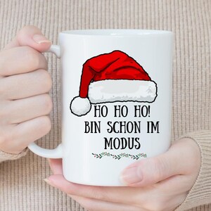 May include: White ceramic mug with a red Santa hat design and the text "HO HO HO! BIN SCHON IM MODUS" in black. The mug has a white handle and a festive green and red border.