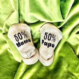 Könnte beinhalten: Ein Paar beige Baby-Socken mit schwarzem Text. Eine Socke sagt "50% Mama" und die andere sagt "50% Papa".