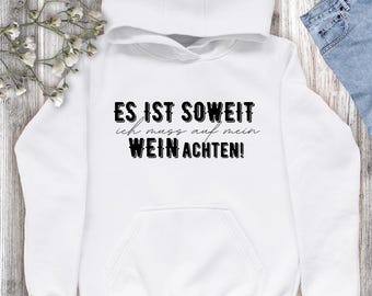 Peut inclure: Sweat à capuche blanc avec le texte allemand "ES IST SOWEIT ich muss auf mein WEIN ACHTEN!" en noir. Le sweat-shirt a une poche avant et une capuche à cordon. L'arrière-plan est en bois clair.