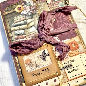 Può includere: Un diario di colore bordeaux e marrone con un'estetica vintage. La copertina presenta un collage di immagini tra cui libri, fiori e un uccello. Il diario è legato con un nastro bordeaux e ha un piccolo occhiello di metallo sulla copertina anteriore. Il testo "No. 46. 313..." è visibile sulla copertina anteriore.