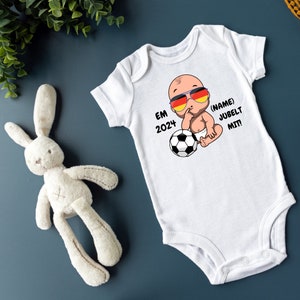 Puede incluir: Un body blanco para bebé con un bebé de dibujos animados que lleva gafas de sol y sostiene un balón de fútbol. El body tiene el texto "EM 2024" y "(NAME) JUBELT MIT!" impreso en él.