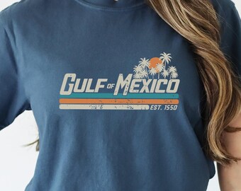 May include: A navy blue t-shirt with a vintage-style graphic that reads "GULF of MEXICO EST. 1950" with palm trees and a sunset.