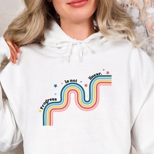 May include: White hoodie with a graphic design of a rainbow-colored wavy line with stars and the text "Progress is not linear."
