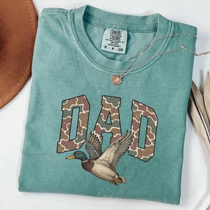 May include: A teal blue t-shirt with a camouflage print "DAD" graphic and a duck illustration. The shirt has a distressed look and is made of a soft, comfortable fabric.