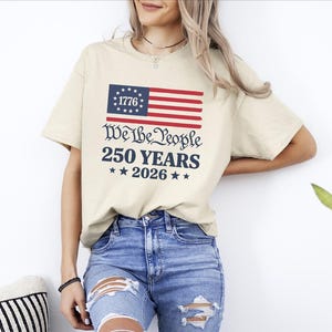 May include: A beige t-shirt featuring an American flag graphic and the text "We the People 250 Years 2026". The shirt is worn with blue jeans. The design incorporates red, white, and blue colours.