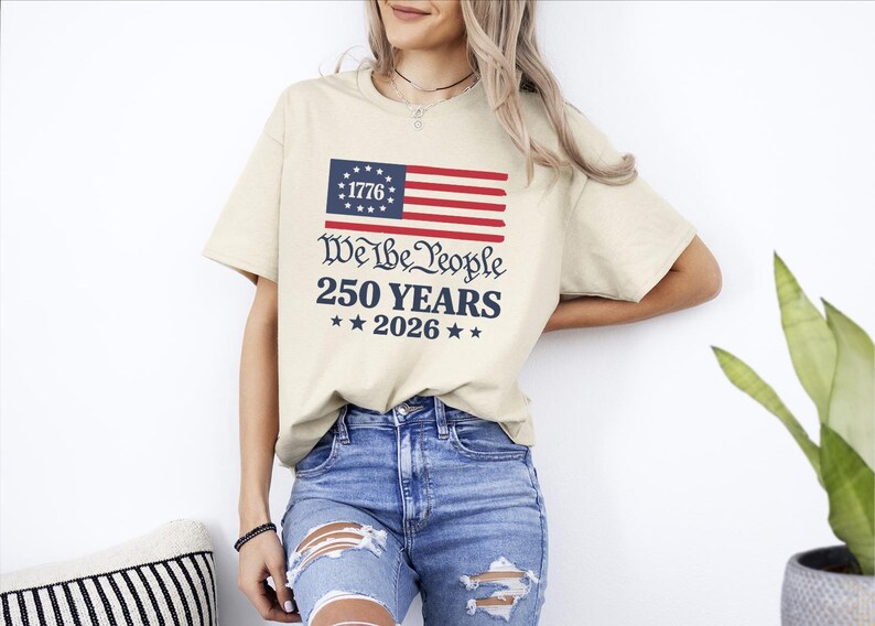 May include: A beige t-shirt featuring an American flag graphic and the text "We the People 250 Years 2026". The shirt is worn with blue jeans. The design incorporates red, white, and blue colours.