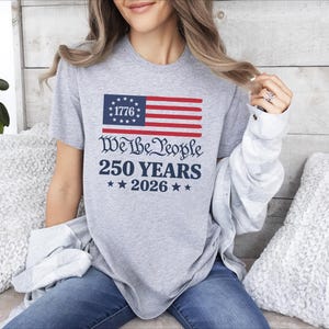 May include: A grey t-shirt with a patriotic design. The design features a stylized American flag with the text "1776" in the blue field, and the words "We the People 250 Years 2026" in a decorative font. The shirt is worn by a person.