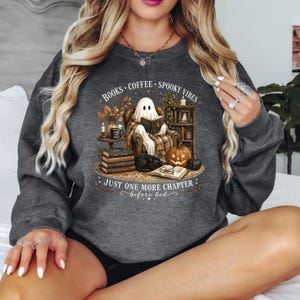May include: A dark gray crewneck sweatshirt featuring a graphic print of a ghost sitting in an armchair reading a book. The design includes a stack of books, a pumpkin, a black cat, and the text: BOOKS, COFFEE, SPOOKY VIBES, JUST ONE MORE CHAPTER before bed.