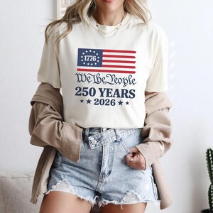 May include: A cream-coloured t-shirt with a patriotic design. The graphic features a stylized American flag with the text "1776" and the words "We the People 250 Years 2026" in navy blue. The shirt is worn with denim shorts.