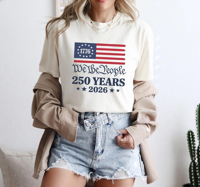 May include: A cream-coloured t-shirt with a patriotic design. The graphic features a stylized American flag with the text "1776" and the words "We the People 250 Years 2026" in navy blue. The shirt is worn with denim shorts.