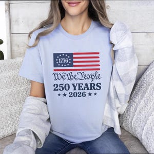 May include: Pale blue t-shirt with a patriotic design. It features a stylized American flag and the text "We the People 250 Years 2026". The flag has a blue rectangle with white stars and red and white stripes. The shirt is a crew neck.