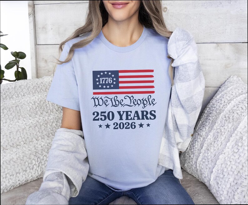 May include: Pale blue t-shirt with a patriotic design. It features a stylized American flag and the text "We the People 250 Years 2026". The flag has a blue rectangle with white stars and red and white stripes. The shirt is a crew neck.