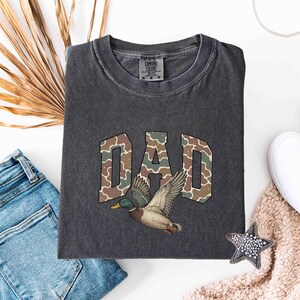 May include: A dark gray t-shirt with a camouflage pattern spelling "DAD" and a duck flying above the word.