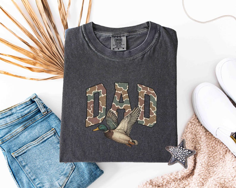 May include: A dark gray t-shirt with a camouflage pattern spelling "DAD" and a duck flying above the word.