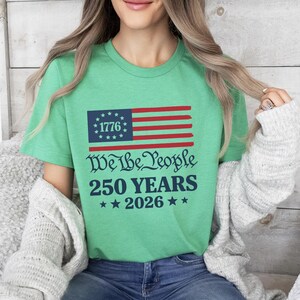 May include: A green t-shirt with a graphic of the American flag, including the text "1776" in a blue box, and red and white stripes. Below the flag are the words "We The People 250 YEARS 2026" in a decorative font, with stars.