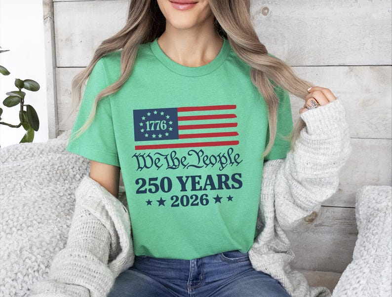 May include: A green t-shirt with a graphic of the American flag, including the text "1776" in a blue box, and red and white stripes. Below the flag are the words "We The People 250 YEARS 2026" in a decorative font, with stars.