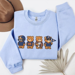 May include: A light blue sweatshirt featuring four cartoon bears dressed in football attire. The bears are wearing helmets, jerseys, and holding a football and a flag. The sweatshirt is a crew neck style.