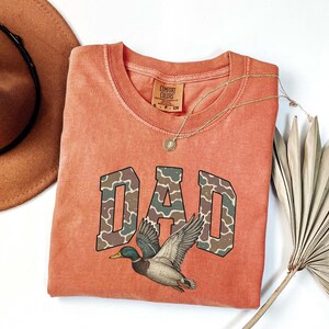 May include: A burnt orange t-shirt with a camouflage print "DAD" graphic and a duck illustration. The shirt has a distressed look and is made by Comfort Colors.