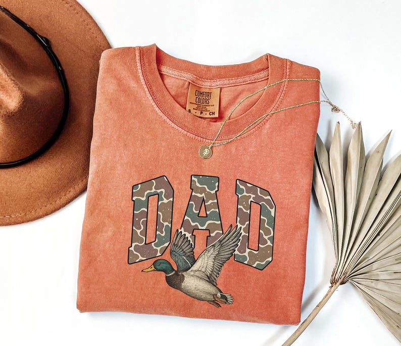 May include: A burnt orange t-shirt with a camouflage print "DAD" graphic and a duck illustration. The shirt has a distressed look and is made by Comfort Colors.