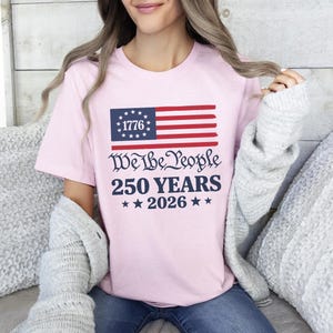May include: A light pink t-shirt with a patriotic design. The design features a stylized American flag with the text "1776" in the blue field, and the words "We the People 250 YEARS 2026" in a dark blue font.