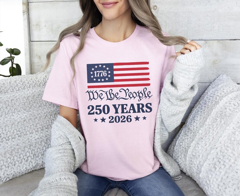 May include: A light pink t-shirt with a patriotic design. The design features a stylized American flag with the text "1776" in the blue field, and the words "We the People 250 YEARS 2026" in a dark blue font.