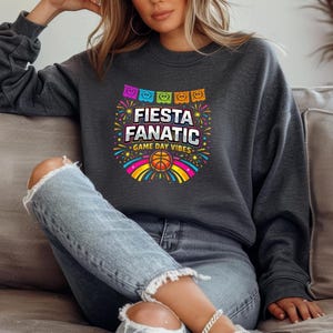 May include: Dark gray sweatshirt with the text "Fiesta Fanatic" in white and yellow, with a basketball graphic. The design includes colorful fireworks, a rainbow, and decorative banners. The sweatshirt is a casual, comfortable style.