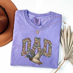 May include: A purple t-shirt with the word "DAD" in camouflage print and a duck flying above it. The shirt is made by Comfort Colors.
