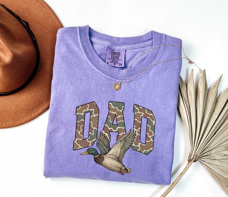 May include: A purple t-shirt with the word "DAD" in camouflage print and a duck flying above it. The shirt is made by Comfort Colors.