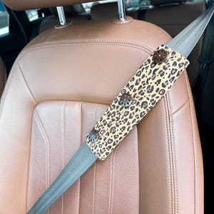 May include: A car seat belt cover with a leopard print design. The cover is tan with brown spots and has three decorative buttons. The seat belt is gray, and the car seat is brown.
