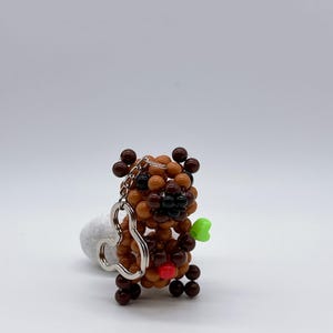 May include: A beaded bear keychain in shades of brown, with a red bead nose and a green bead held in its hand. The keychain has a silver-colored heart-shaped attachment.