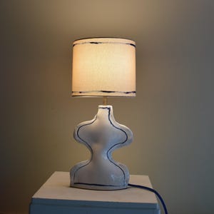 May include: A white ceramic table lamp with a unique, abstract form. The lamp features a cream-colored lampshade with dark blue trim. The base is outlined with dark blue lines. A blue power cord extends from the base.