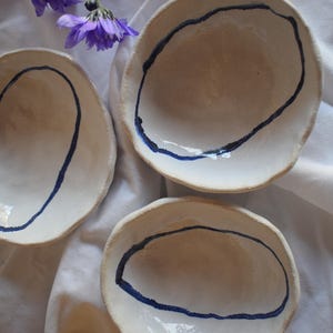 May include: Three handmade white ceramic bowls with a dark blue painted oval design. The bowls are arranged on a white cloth, with purple flowers in the background. The bowls have a rustic, organic shape.