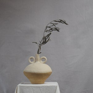 以下が含まれることがあります： 2つの取っ手と底に茶色の釉薬が施された白い陶器の花瓶。花瓶にはオリーブの葉の乾燥した枝が挿されています。