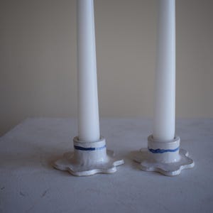 May include: Two white taper candles in ceramic candle holders. The candle holders are white with a blue stripe and a scalloped edge. The candles are centered on a white surface.