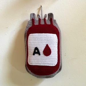 May include: A crocheted blood bag with a white label that says 'A' and has a red blood drop. The bag is red and gray.