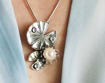 May include: A silver necklace with a pendant featuring a stylized lily pad design with a pearl and blue gemstones. The pendant is on a silver chain.