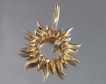 May include: A gold-colored pendant in the shape of a sunburst, featuring a central open circle and radiating petal-like elements. A small, round diamond is set near the edge. The Etsy shop name "wingedlion.etsy.com" is at the bottom.