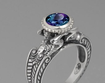 May include: A silver ring with an oval, color-changing gemstone. The ring features intricate, detailed carvings and a beaded bezel setting around the stone. The band has a decorative pattern. The website address "wingedlion.etsy.com" is visible.