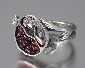 May include: A silver ring with a pomegranate design. The pomegranate is open and contains a woman figure and red gemstones. The ring band is textured and has a leaf design.