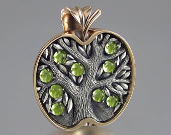May include: A silver pendant shaped like an apple with a gold stem and leaves. The inside of the apple features a detailed tree with green peridot gemstones.