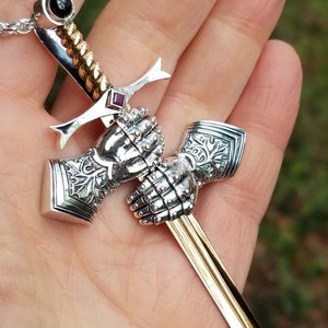 May include: A silver pendant necklace featuring a miniature sword design. The sword has a gold-colored blade and a handle with intricate detailing. The necklace includes a black stone and a red stone. The text "wingedlion.etsy.com" is visible.