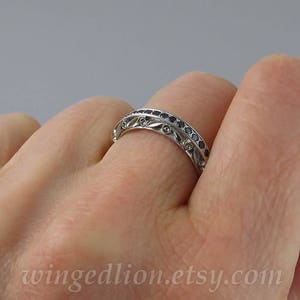 Op de afbeelding: Een zilveren bandring met een decoratief ontwerp met kleine zwarte edelstenen. De ring wordt aan een vinger gedragen.