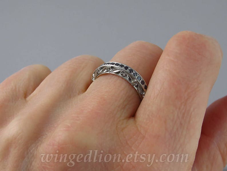 Op de afbeelding: Een zilveren bandring met een decoratief ontwerp met kleine zwarte edelstenen. De ring wordt aan een vinger gedragen.