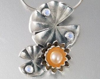 May include: A silver pendant necklace featuring a stylized flower with a peach-colored center and a pearl. The flower is surrounded by two silver leaves with moonstone accents.