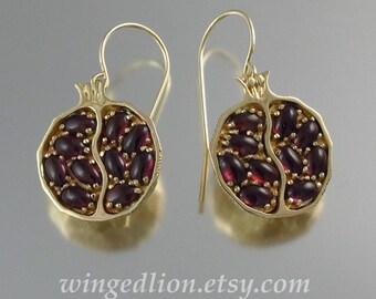 May include: A pair of gold drop earrings shaped like halved pomegranates. Each earring features a textured gold frame filled with small, oval-shaped, deep red garnet gemstones arranged to resemble pomegranate seeds. The text wingedlion.etsy.com is visible.