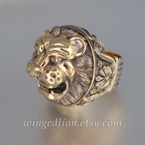 Può includere: Un anello in oro a forma di testa di leone con occhi di pietre preziose rosse. La fascia dell'anello è decorata con motivi floreali.