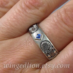 May include: A silver band ring with a blue gemstone set in a square bezel. The ring is decorated with an intricate floral design.